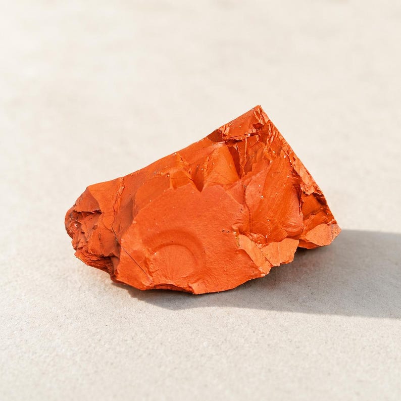 May include: A raw, unpolished piece of red jasper stone with a vibrant orange-red hue. The mineral specimen features a jagged, angular shape with a matte texture and visible conchoidal fractures across its surface, showcasing natural earthy tones.