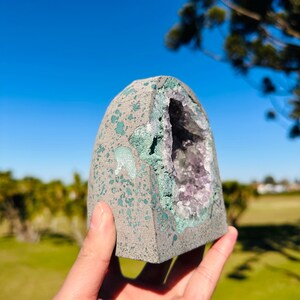 May include: A dome-shaped geode with a rough, gray exterior speckled with light green paint. The center features a hollow cavity lined with sparkling purple amethyst crystals, framed by a jagged, textured edge of green mineral deposits.