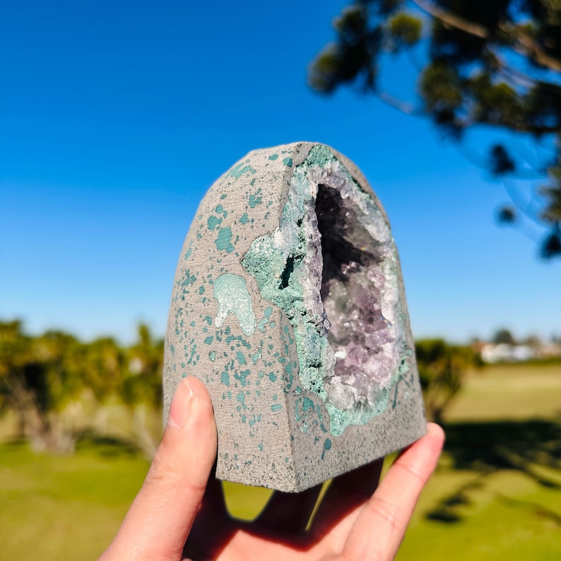 May include: A dome-shaped geode with a rough, gray exterior speckled with light green paint. The center features a hollow cavity lined with sparkling purple amethyst crystals, framed by a jagged, textured edge of green mineral deposits.