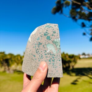May include: A semi-circular stone specimen featuring a textured grey base with scattered, irregular patches of vibrant teal and light green mineral inclusions across its flat, polished surface, held against a clear blue sky background.