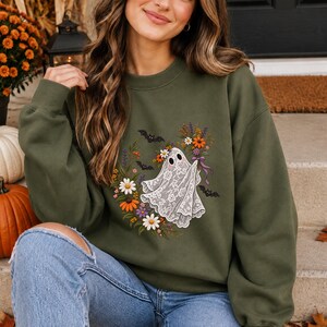 Puede incluir: Una sudadera de cuello redondo color verde oliva con un gráfico de un fantasma con patrón de encaje blanco que sostiene un ramo de flores otoñales coloridas. El diseño está rodeado por una corona floral y pequeños murciélagos negros.