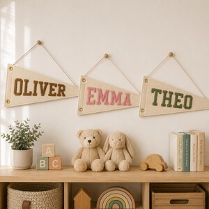 May include: Three triangular cream-colored felt pennants hanging on a wall, each featuring a name in block letters: OLIVER in brown, EMMA in pink, and THEO in green. Each pennant is suspended by a thin string attached to a small wooden peg.
