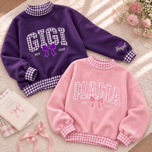 Könnte beinhalten: Ein lila Sweatshirt mit lila Vichy-Karo-Kragen, Bündchen und Saum, mit dem Text GIGI, einer Schleife und EST 2026, sowie ein rosa Sweatshirt mit rosa Vichy-Karo-Kragen, Bündchen und Saum, mit dem Text MAMA, einer Schleife und EST 2026.