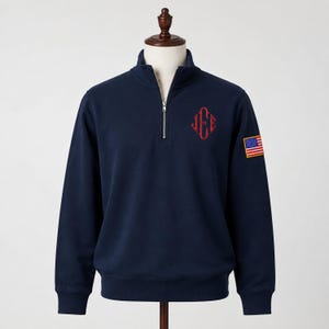 May include: A navy blue quarter-zip pullover featuring a red embroidered monogram with the letters JEE on the left chest and a small rectangular American flag patch on the left sleeve. The garment has a stand-up collar and a silver metal zipper.