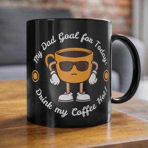 Könnte beinhalten: Schwarze Keramik-Kaffeetasse mit einem Cartoon-Kaffeetassen-Design, das eine Sonnenbrille und Handschuhe trägt. Der Text lautet: "Mein Papa-Ziel für heute: Trinke meinen Kaffee heiß!" Ein lustiges Geschenk.