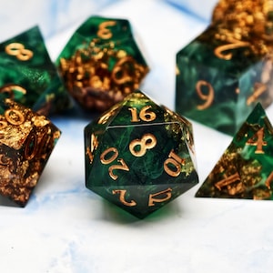 Puede incluir: Un conjunto de dados poliédricos de varias formas, incluido un dado de veinte caras. Los dados son de color verde oscuro con números dorados y motas doradas incrustadas en el material translúcido. Dados para juegos de mesa.