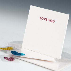 May include: A white card with the words "LOVE YOU" printed in red. The card is propped up on a white envelope with three heart-shaped stickers on the envelope. The stickers are yellow, blue, and pink.