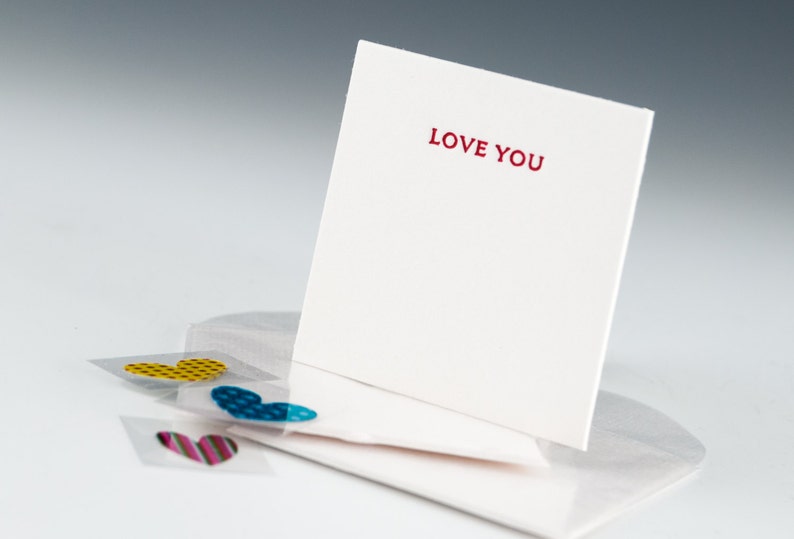 May include: A white card with the words "LOVE YOU" printed in red. The card is propped up on a white envelope with three heart-shaped stickers on the envelope. The stickers are yellow, blue, and pink.