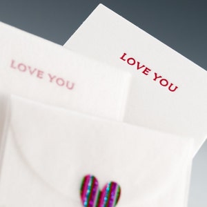 May include: Two white envelopes with the words "Love You" printed in red on each. The top envelope is folded over the bottom envelope. A sparkly pink and blue heart is on the bottom envelope.