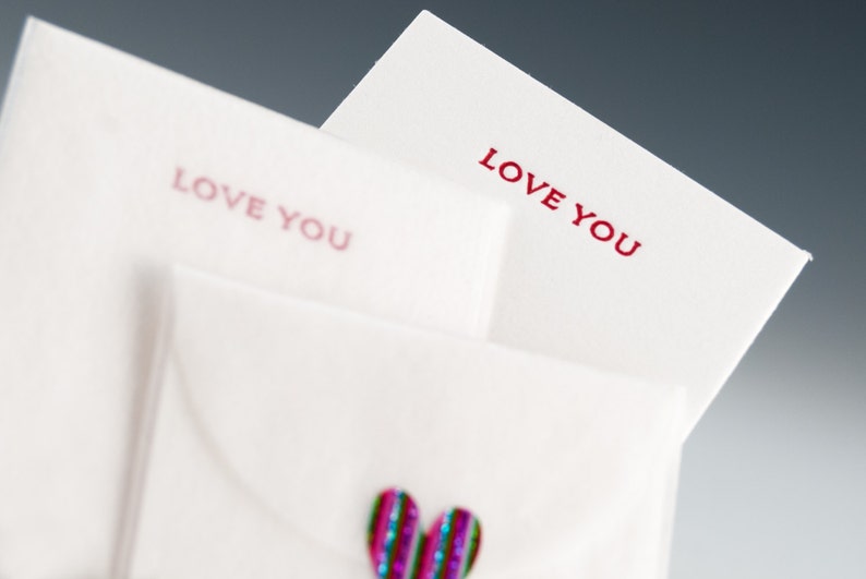 May include: Two white envelopes with the words "Love You" printed in red on each. The top envelope is folded over the bottom envelope. A sparkly pink and blue heart is on the bottom envelope.