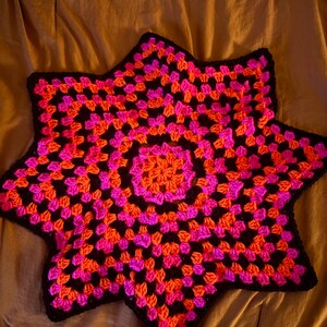 May include: A handmade, star-shaped crochet item in vibrant colors. The design features a central orange circle surrounded by concentric rings of pink and orange squares, all outlined in black. This decorative piece is likely a pillow or a blanket.