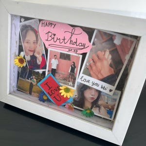 Op de afbeelding: Een witte schaduwdoos met meerdere foto's, decoratieve bloemen en handgeschreven tekst. De tekst bevat "Happy Birthday" op een roze hartvormige uitsparing en "I love U" op een rode kaart. Foto's tonen mensen en handen.