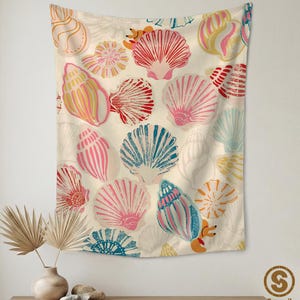 May include: A rectangular wall tapestry featuring a repeating pattern of various seashells in shades of pink, red, blue, and yellow on a cream background. The design includes fan-shaped, spiral, and conch shells with textured line details.