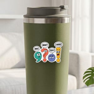 Op de afbeelding: Een olijfgroene reisbeker met een grijze deksel en een sticker van vier kleurrijke leestekens met gezichten en tekstballonnen: WAIT, WHAT?, STOP IT. en I AGREE!. De karakters zijn een groenblauwe komma, een rood vraagteken, een blauwe punt en een geel uitroepteken.