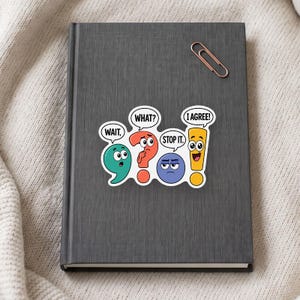 Op de afbeelding: Een donkergrijs notitieboek met een koperkleurige paperclip en een sticker met vier antropomorfe leestekens met tekstballonnen die de tekst: WAIT, WHAT?, STOP IT. en I AGREE! in zwarte hoofdletters bevatten.