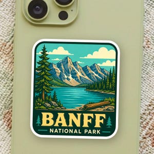 May include: A square sticker with rounded corners featuring a landscape illustration of Banff National Park. The design shows a blue lake, snow-capped mountains, pine trees, and a log on the shore. The text BANFF NATIONAL PARK is printed in yellow.