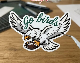 Op de afbeelding: Een gestanste sticker met een witte en grijze adelaar met een gele snavel en klauwen, die een bruine football vasthoudt. Boven de adelaar staat de tekst Go birds in een donkergroen cursief lettertype. De sticker heeft een witte rand.