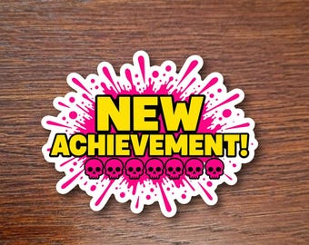 May include: A die-cut sticker featuring a bright pink ink splatter design with a white border. The text NEW ACHIEVEMENT! is printed in bold yellow letters. Below the text, seven small pink skull icons are arranged in a horizontal row.