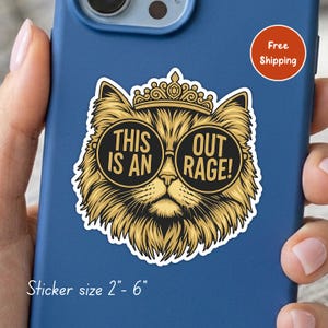 May include: A die-cut sticker featuring a golden-furred cat wearing a crown and black circular sunglasses. The text on the lenses reads THIS IS AN OUT RAGE!. The sticker is applied to a blue phone case. The sticker size is listed as 2"- 6".
