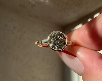May include: A gold ring featuring a large round brilliant-cut center stone set in a bezel with milgrain detailing. The ring is accented by two tapered baguette-cut side stones, one on each side of the central setting, mounted on a gold band.