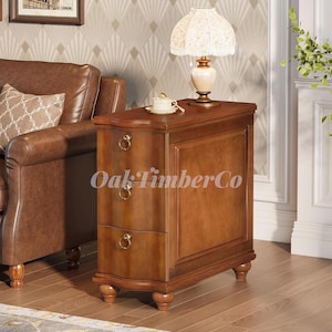 May include: A wooden side table with two drawers and a cabinet door. The table is a warm brown color with brass hardware and a curved base. A lamp with a patterned shade sits on top. The text "Oak Timber Co" is visible.