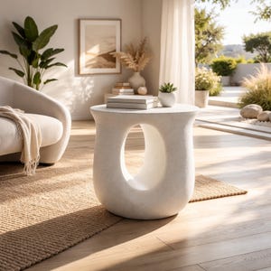 May include: A white, modern side table with a unique, open oval design. The table is made of a textured material and has a round top. A small succulent in a white pot and a stack of books sit on top. The table is 18 inches tall.