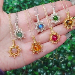 Könnte beinhalten: Eine Sammlung von Sonnenanhänger-Halsketten mit verschiedenen Edelsteinzentren. Die Halsketten haben goldene und silberne Ketten und drahtgewickelte Fassungen. Zu den Edelsteinen gehören rosa, orange, grüne und blaue Steine.