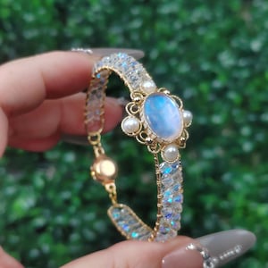 May include: A gold-toned bracelet featuring a large, oval, blue moonstone center stone, surrounded by small pearls and intricate gold filigree. The bracelet band is made of clear, iridescent beads. A gold clasp secures the bracelet.
