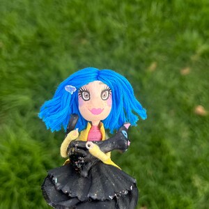 May include: A handmade doll with blue hair, large eyes, and a pink mouth. The doll wears a black dress, yellow jacket, and holds a black cat. The doll's legs are striped pink and white. The background is blurred green grass.