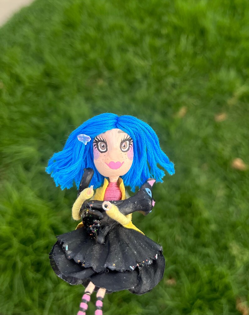May include: A handmade doll with blue hair, large eyes, and a pink mouth. The doll wears a black dress, yellow jacket, and holds a black cat. The doll's legs are striped pink and white. The background is blurred green grass.