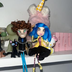 May include: Handmade dolls with detailed features, including blue and brown hair, and black outfits. One doll wears a yellow coat and black skirt. A black cat figurine and a pink Hello Kitty are also visible.