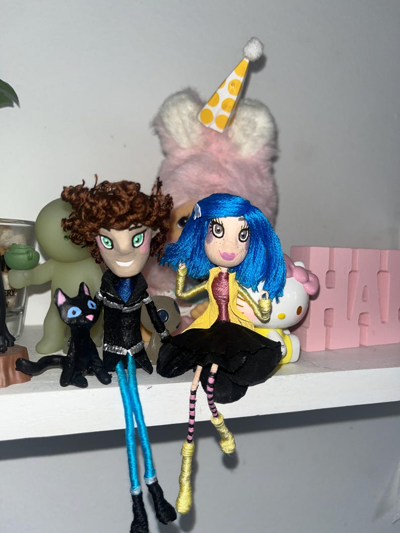 May include: Handmade dolls with detailed features, including blue and brown hair, and black outfits. One doll wears a yellow coat and black skirt. A black cat figurine and a pink Hello Kitty are also visible.