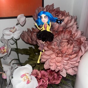 May include: A handmade doll with blue hair, a yellow jacket, and a black skirt, perched atop a large, pink flower arrangement. A small black cat figure is next to the doll. White orchids and other flowers are in the background.