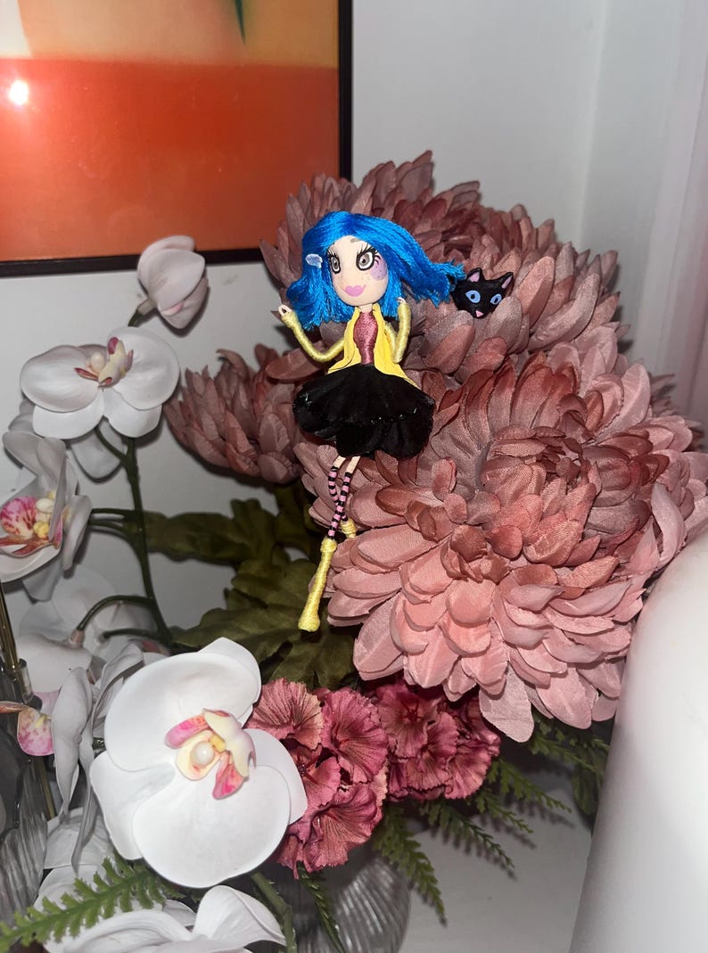May include: A handmade doll with blue hair, a yellow jacket, and a black skirt, perched atop a large, pink flower arrangement. A small black cat figure is next to the doll. White orchids and other flowers are in the background.