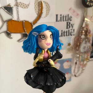 May include: A handmade doll with blue hair, a pink top, and a black skirt. The doll has a yellow jacket and striped legs. It is holding a black object. The doll is a whimsical, artistic creation.