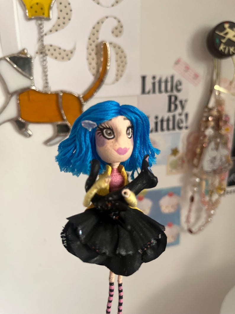 May include: A handmade doll with blue hair, a pink top, and a black skirt. The doll has a yellow jacket and striped legs. It is holding a black object. The doll is a whimsical, artistic creation.
