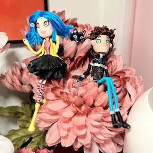 May include: Handmade doll figures of a girl with blue hair and a boy with brown hair, each with detailed clothing. A black cat figure is between them. The dolls are set on a bed of pink flowers, creating a whimsical scene.