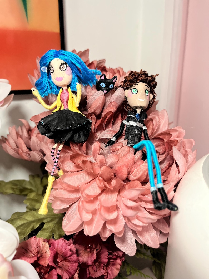 May include: Handmade doll figures of a girl with blue hair and a boy with brown hair, each with detailed clothing. A black cat figure is between them. The dolls are set on a bed of pink flowers, creating a whimsical scene.