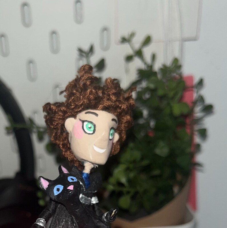 May include: A handmade figurine of a character with brown curly hair, green eyes, and pink cheeks. The figure is wearing a black coat and holding a black cat with blue eyes. A small plant is in the background.