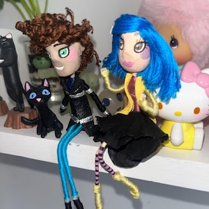 May include: Two handmade dolls, one with brown hair and a black outfit, the other with blue hair and a yellow jacket. Both dolls have painted faces and yarn legs. A black cat figurine and a Hello Kitty figure are in the background.
