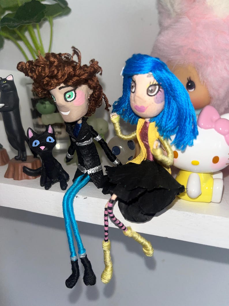 May include: Two handmade dolls, one with brown hair and a black outfit, the other with blue hair and a yellow jacket. Both dolls have painted faces and yarn legs. A black cat figurine and a Hello Kitty figure are in the background.