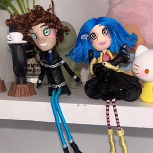 May include: Two handmade dolls with unique features. One doll has brown curly hair, green eyes, and a black coat with blue legs. The other doll has blue hair, a black dress, and holds a black cat. Both dolls have painted faces.