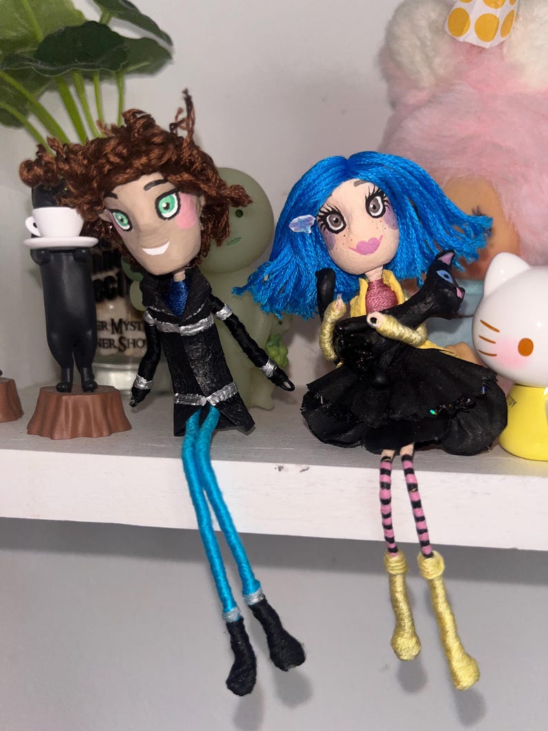 May include: Two handmade dolls with unique features. One doll has brown curly hair, green eyes, and a black coat with blue legs. The other doll has blue hair, a black dress, and holds a black cat. Both dolls have painted faces.