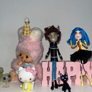 May include: A collection of figurines and toys, including a pink bunny with a party hat, a Hello Kitty figure, and a doll with blue hair. A pink block spells out the word "HAPPY". A black cat figurine is also present.