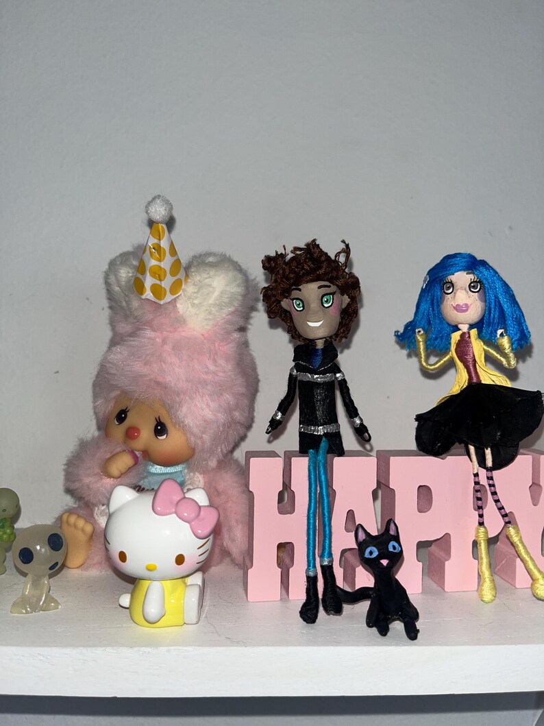 May include: A collection of figurines and toys, including a pink bunny with a party hat, a Hello Kitty figure, and a doll with blue hair. A pink block spells out the word "HAPPY". A black cat figurine is also present.