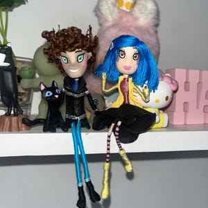 May include: Handmade dolls of characters with detailed features. One doll has brown hair, a black coat, and blue pants. The other doll has blue hair, a yellow coat, and a black skirt. Both dolls have painted faces and are posed sitting on a shelf.