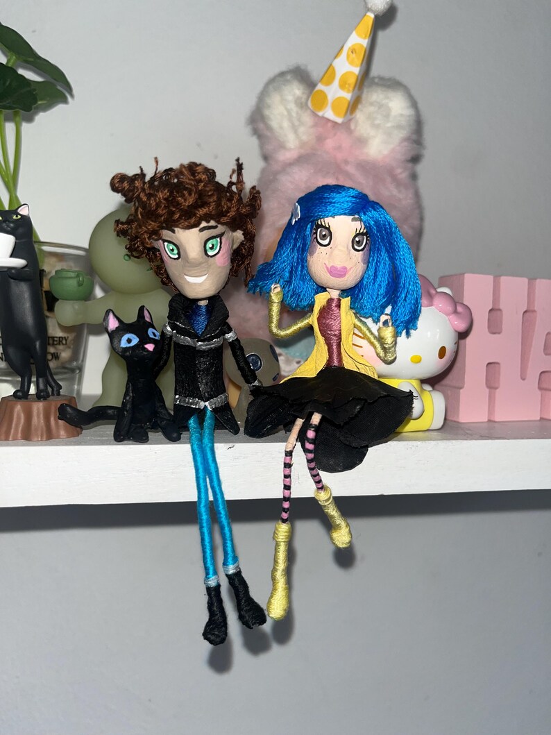 May include: Handmade dolls of characters with detailed features. One doll has brown hair, a black coat, and blue pants. The other doll has blue hair, a yellow coat, and a black skirt. Both dolls have painted faces and are posed sitting on a shelf.