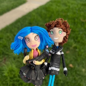 May include: Handmade dolls of a girl with blue hair and a boy with brown hair. The girl doll wears a black dress and holds a black cat. The boy doll wears a black jacket with white stripes. Both dolls have blue legs.