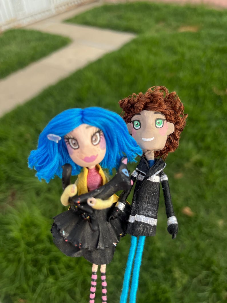 May include: Handmade dolls of a girl with blue hair and a boy with brown hair. The girl doll wears a black dress and holds a black cat. The boy doll wears a black jacket with white stripes. Both dolls have blue legs.