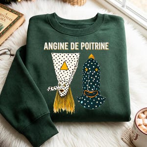 May include: A dark green crewneck sweatshirt featuring the text "ANGINE DE POITRINE" in white letters. Below the text are two stylized figures: one with a white polka-dot cone head and yellow hair, and another with a dark blue polka-dot head.